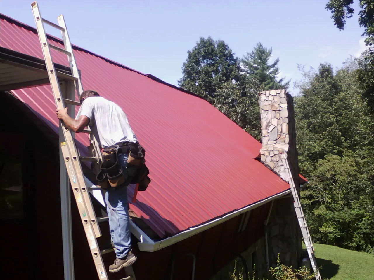 Roofer on a ladder beside a red metal roof and stone chimney — Soffit & Fascia Repair by Roofing Brothers in Eureka